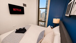 A hotel room with a bed, a TV displaying 'NETFLIX,' a window with a city view, and a lamp on the nightstand. - 25th Floor 2BR Grant Park Suite with Balcony (Chicago)