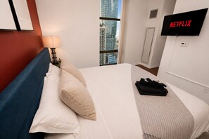 A hotel room with a bed, a TV mounted on the wall, a window with a view of buildings, and a lamp on the nightstand.