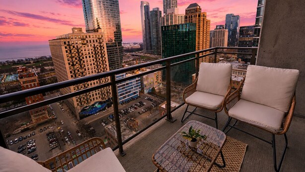 A balcony with two wicker chairs and a small table, overlooking a cityscape with tall buildings and a sunset sky. - 25th Floor 2BR Grant Park Suite with Balcony (Chicago)