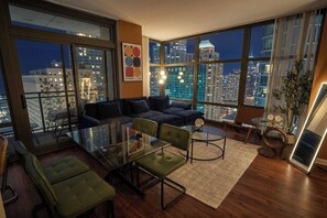 En moderne stue med et glasbord, grønne stole, en mørk sofa og et stort vindue med udsigt over byen.