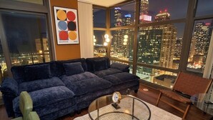 A modern living room with a large window offering a city view, a dark blue sofa, a round glass coffee table, and a wooden bench. - 25th Floor 2BR Grant Park Suite with Balcony (Chicago)