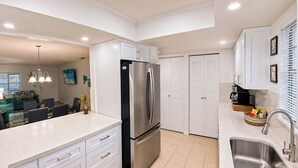 A modern kitchen with white cabinets, a stainless steel refrigerator, a sink, and a dining area with a green couch and a chandelier. - Stylish 2BR/2BA coastal getaway to 7 Min Drive to Beach "Crystal Bay Stays" (Englewood)