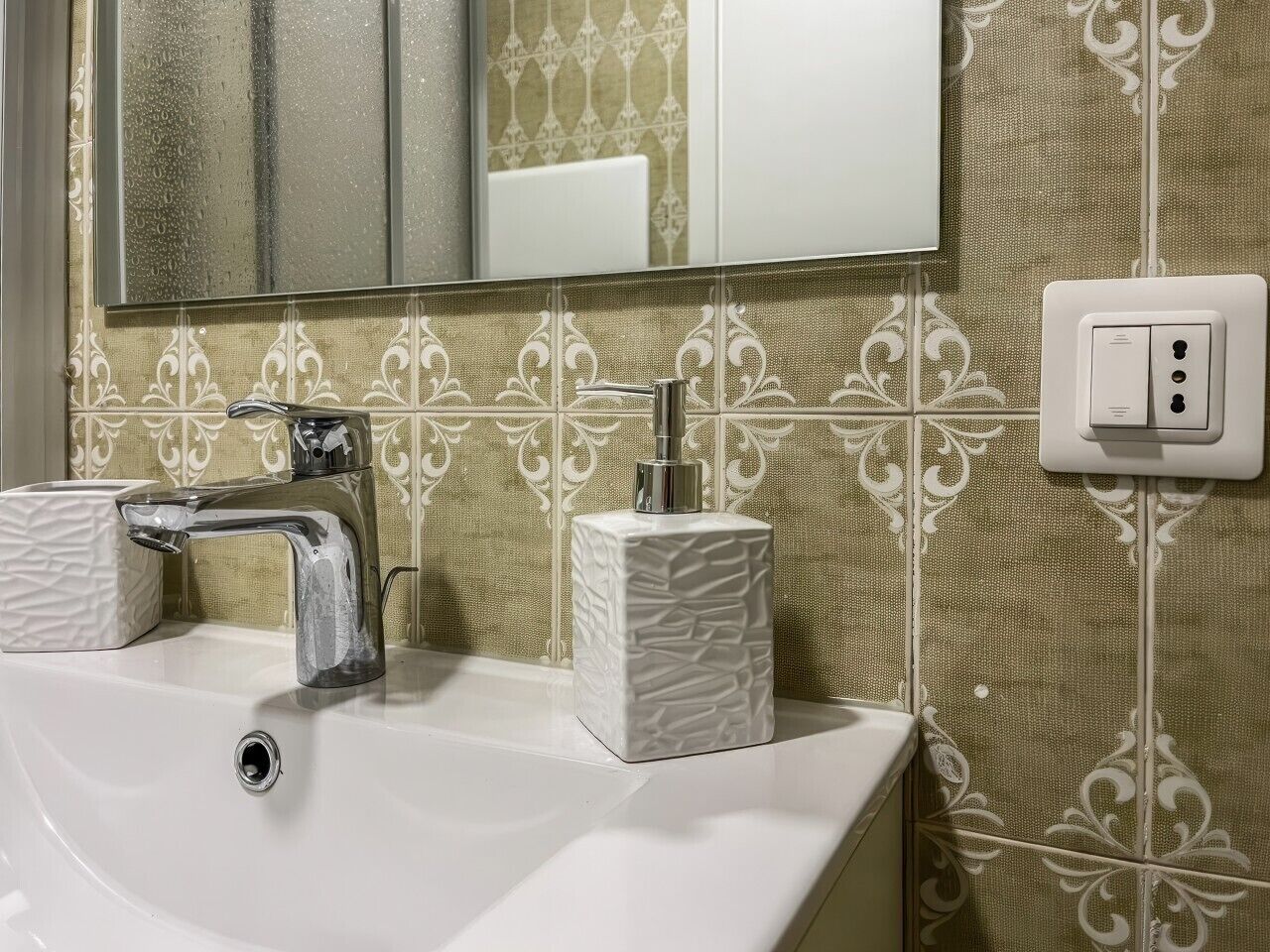 Een badkamer met een wit wastafel, een chromen kraan en een spiegel erboven. De wand is versierd met een patroon en er zit een witte stopcontact in de muur.