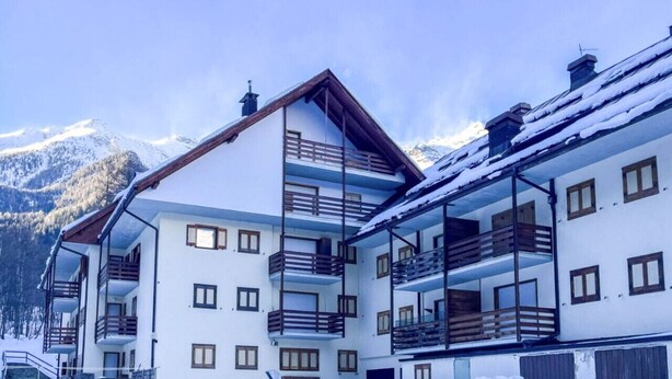 A multi-story building with balconies, a snow-covered roof, and a mountainous backdrop. - Casa Castoro - In the heart of Val di Peio (Peio Fonti)