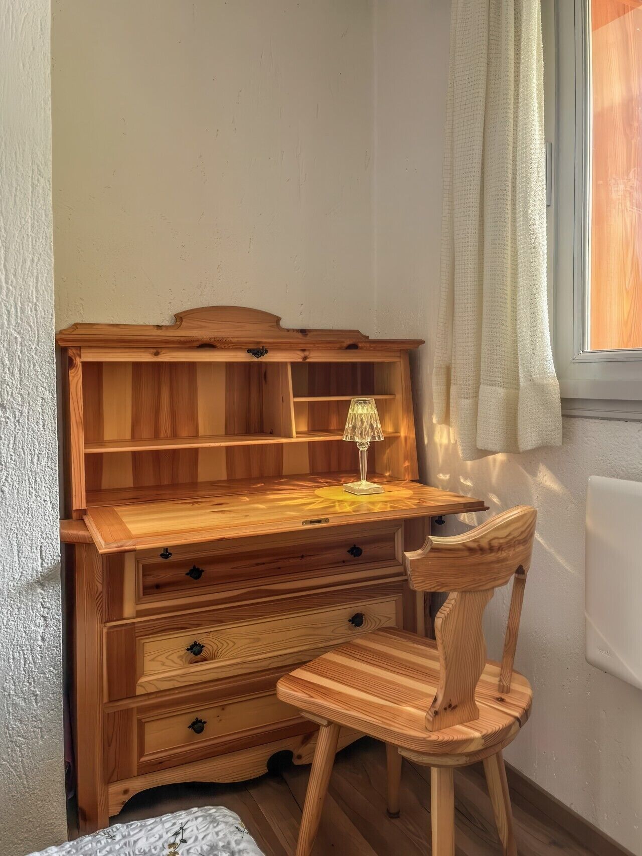 Uma escrivaninha de madeira com gavetas e uma lâmpada, uma cadeira e uma janela com cortinas.