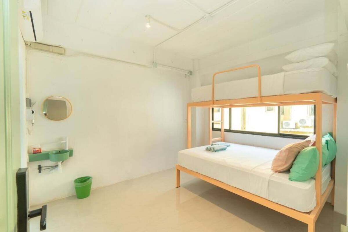 Una cama individual superpuesta con ventana, un lavamanos y un cubo de basura verde en una habitación con paredes blancas.