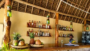A rustic bar area with wooden shelves stocked with various liquor bottles, a wooden counter, and tropical fruits displayed on the counter. - Moayana Liora Zanzibar · Pongwe · Beachfront - Authentic (Zanzibar)