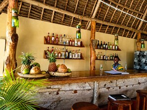 Angolo bar rustico con scaffali in legno pieni di bottiglie di liquori, un bancone di legno e frutta tropicale esposta.