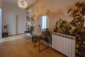 A room with a wooden floor, two wicker chairs, a round mirror, a radiator, and a wall mural of trees.