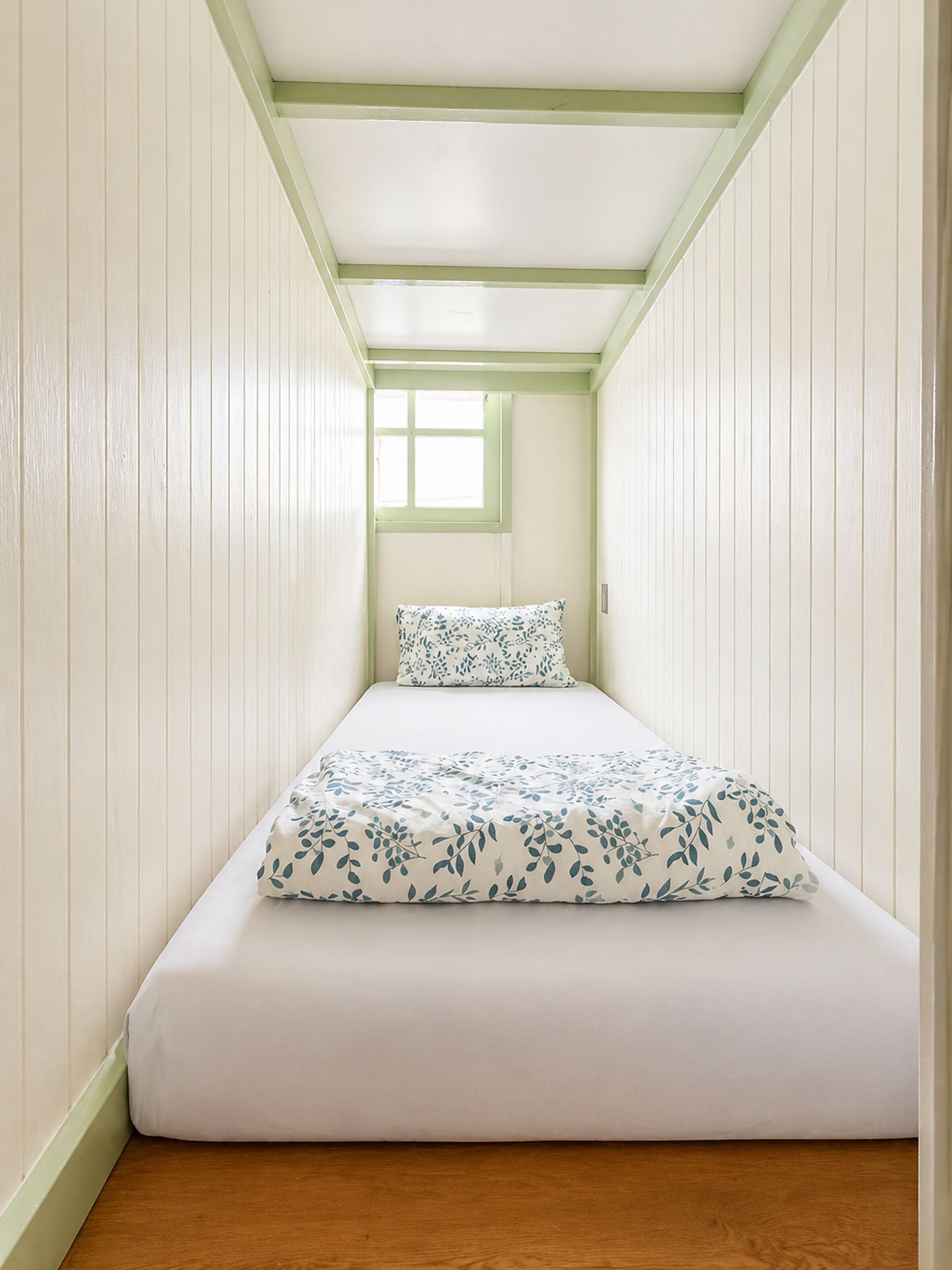 A narrow room with a bed, a window, and wooden flooring.