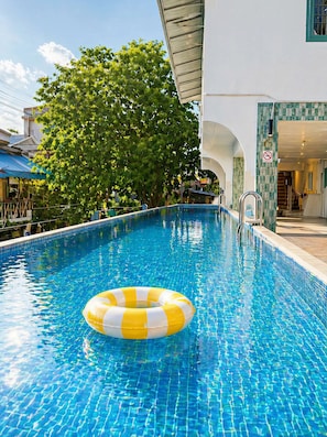 Una piscina con un aro inflable amarillo y blanco, rodeada por un edificio con fachada de azulejos y un gran árbol.