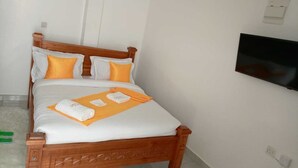 A single bed with white and orange bedding, a wooden headboard, and a flat-screen TV mounted on the wall. - Ehtans Hotel And Accommodations (Eldoret)