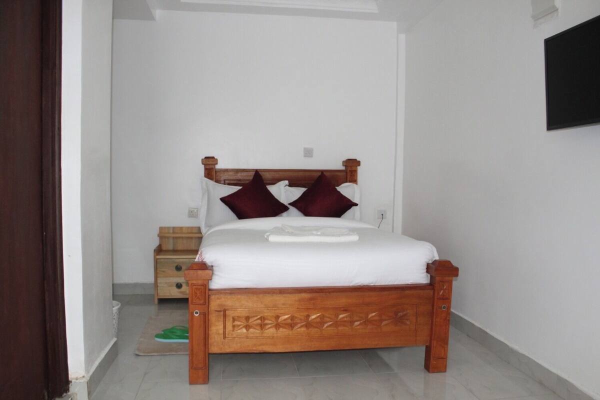 A bedroom with a wooden bed, white bedding, two red pillows, a bedside table, and a green mat.