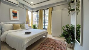 Une chambre avec un lit, un appareil de climatisation, un ventilateur de plafond, de grandes fenêtres avec des rideaux, un tapis coloré et une plante en pot.
