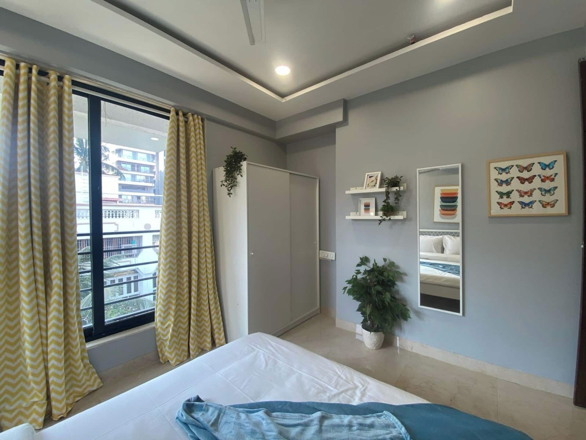 A bedroom with a bed, curtains, a potted plant, a mirror, and wall shelves.
