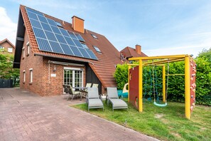 Ett hus med ett sluttande tak, solpaneler, en fasad av tegel, en uteplats med stolar och en lekplats för barn.