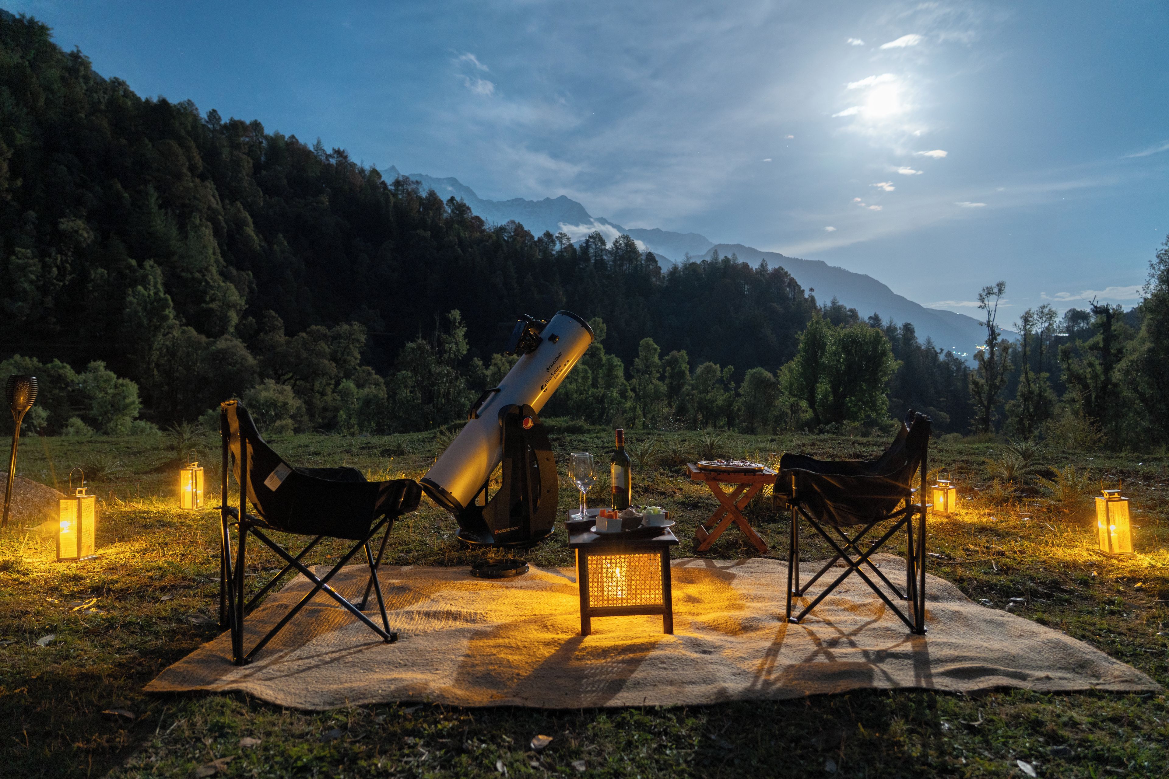An outdoor telescope setup on a blanket with chairs, surrounded by lanterns and a bottle, with mountains in the background.