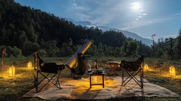 An outdoor telescope setup on a blanket with chairs, surrounded by lanterns and a bottle, with mountains in the background.