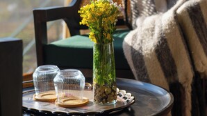 Una mesa con un jarrón de flores, vasos transparentes y una bandeja. Una silla con una manta a rayas se ve al fondo.