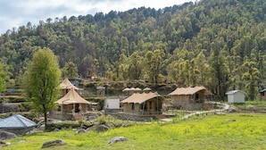 Entorno rural con varias tiendas de campaña y un edificio pequeño, rodeado de un paisaje verde exuberante y una colina boscosa al fondo.
