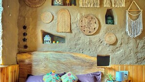 A rustic room with a bed, a fan, and decorative woven items on the walls. - Cozy, Aesthetic Nature Retreat: Feel Your Soul in the Village Soil (Soil)