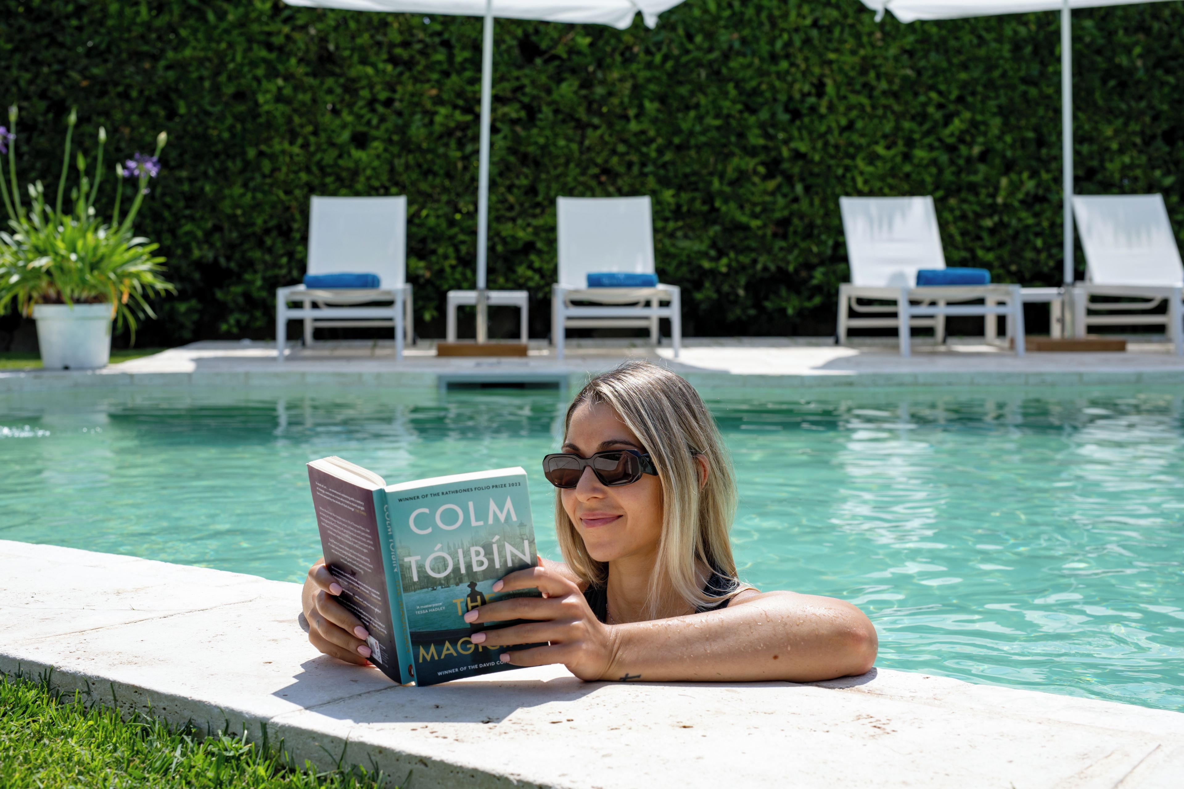 Una persona leyendo un libro de Colm Tóibín junto a una piscina con sillones de descanso y una siega al fondo.