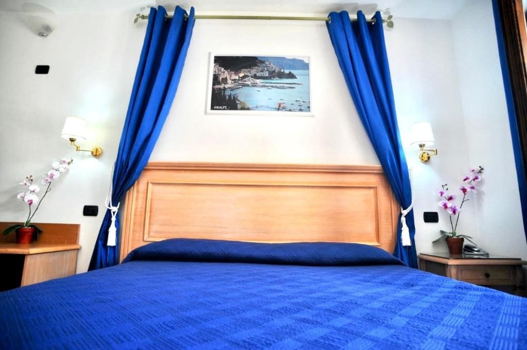 Una habitación de hotel con una cama grande, cortinas azules, un cuadro en la pared y dos mesitas de noche con lámparas.