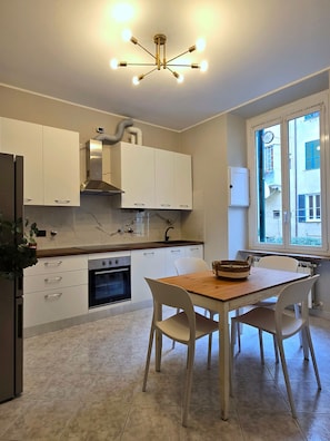 A modern kitchen with white cabinets, a built-in oven, a dining table with chairs, and a window with a view of buildings.
