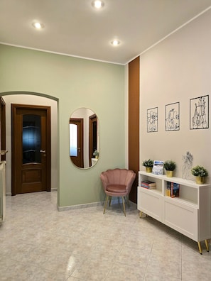 A hallway with a wooden door, a round mirror, a pink chair, a white console table with potted plants, and framed artwork on the wall.