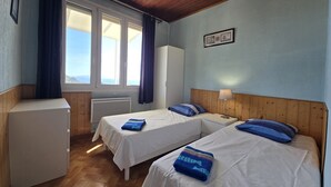 Habitació amb dos llits, un panell de fusta al capçal, un armari blanc, un radiador i una finestra amb cortines.