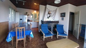 Zona de menjador amb taula i cadires, cuina al fons i una llar de foc amb objectes decoratius.