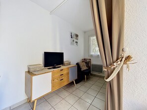 Una habitación con televisión sobre un mueble de madera, un radiador, una ventana con cortinas y una pared decorada con cuadros.