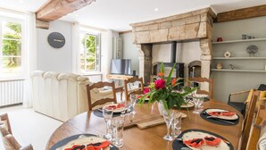 A dining area with a round table set for four, a fireplace, and a television in the background. - Beautiful stone house, private garden in the heart of Cotentin (Bricquebec-en-Cotentin)
