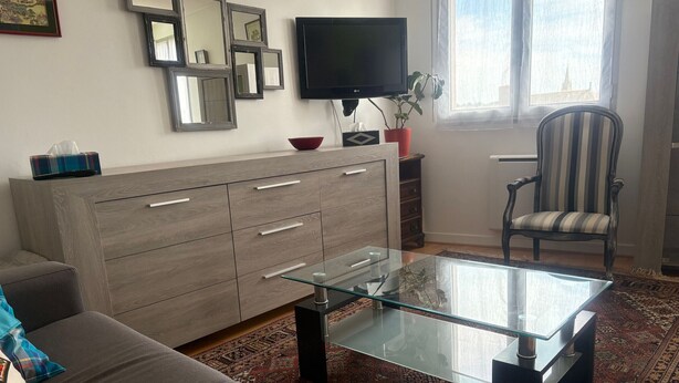 A living room with a sofa, a glass coffee table, a TV, a cabinet, a chair, and a window with a view. - Chic city center studio, parking - 2 guests (Sablé-sur-Sarthe)