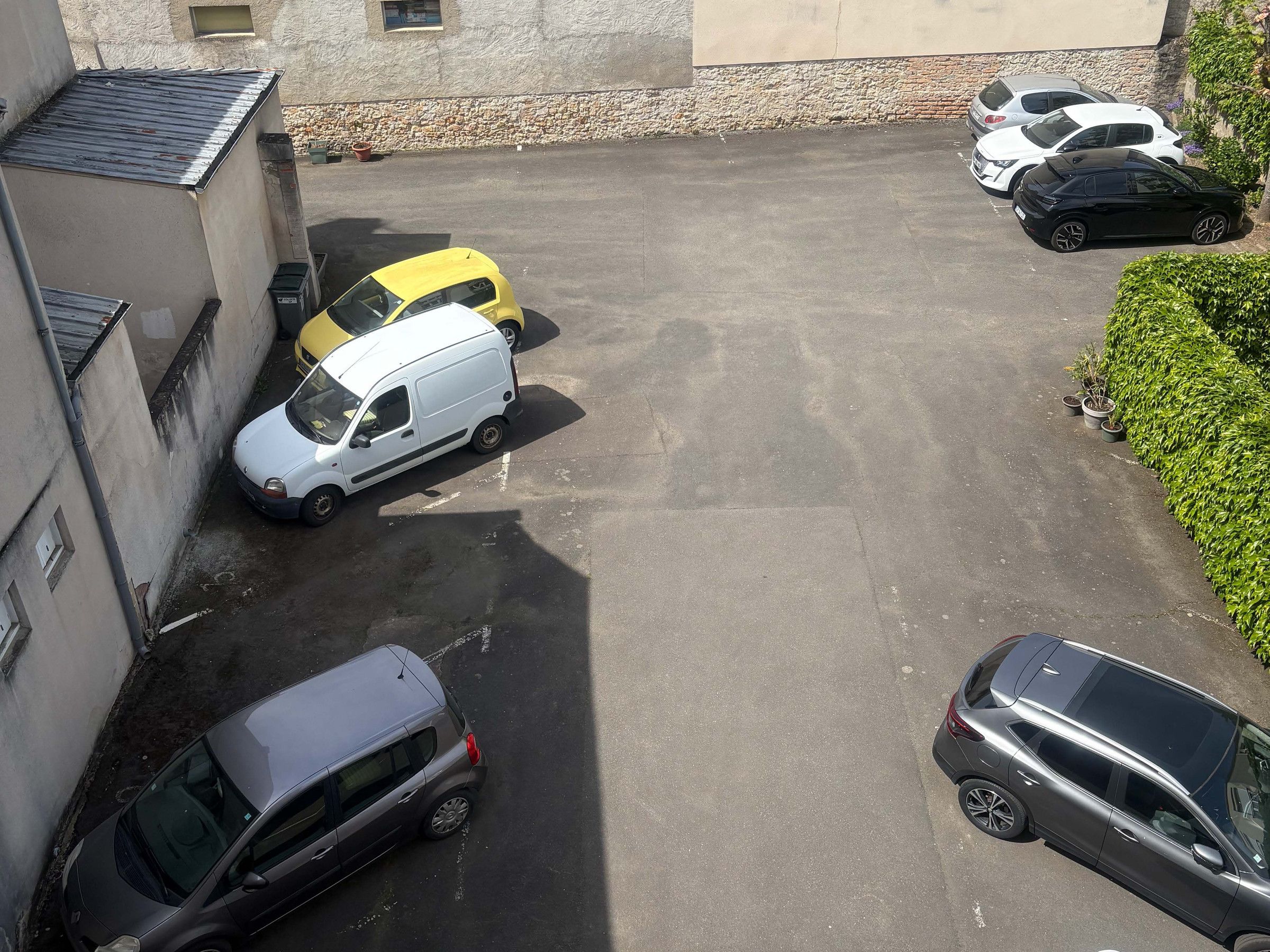 Uma área de estacionamento com vários carros, um edifício de fachada em pedra e um conjunto de arbustos.