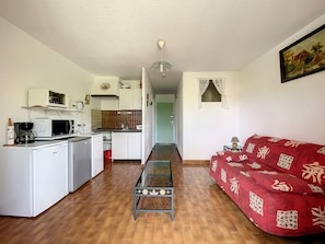 Eine kompakte Küche mit Mikrowelle, Kaffeemaschine und kleinem Kühlschrank. Ein Glastisch befindet sich in der Mitte, und eine rote Couch steht an der Wand.