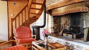 Ein gemütliches Zimmer mit Kamin aus Stein, einer Holztreppe, Korbmöbeln und einem Holztisch mit einer Vase.