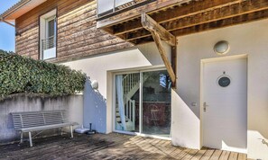 Une maison en bois avec un balcon, un banc, une porte-fenêtre coulissante et une porte blanche avec une plaque numérotée.