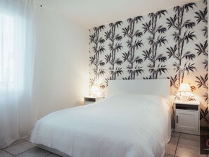 Ein Schlafzimmer mit einem Bett, zwei Nachttischen mit Lampen, einer Wand mit Bambusmuster und einem Fenster mit durchsichtigen Vorhängen.