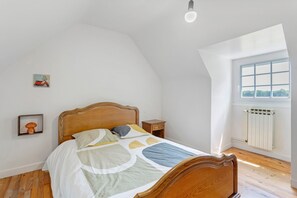 Une chambre avec un lit en bois, une table de chevet, une étagère murale ornée d’un champignon, une fenêtre donnant sur un paysage et un radiateur.