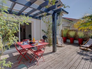 Eine Holzterrasse mit einem roten Tisch, Stühlen, einem Grill und Kübelpflanzen.
