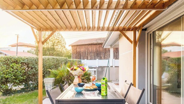 Eine Außenpatio mit einem für das Frühstück gedeckten Tisch, einer hölzernen Pergola darüber und Blick auf eine Wohngegend.
