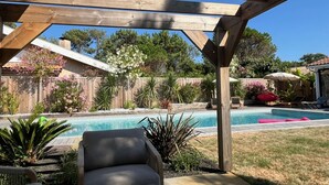 Een houten pergola boven een zwembad in de achtertuin, met een stoel en ligbedden, omgeven door diverse planten en een houten schutting.