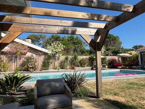 Eine Holz-Pergola über einem Pool im Garten mit einem Stuhl und Liegestühlen, umgeben von Pflanzen und einem Holzzaun.