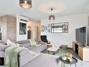 Un salon moderne avec un canapé gris, un fauteuil noir, une petite table basse, un téléviseur et un coin cuisine doté de meubles en bois.