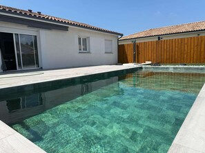 A clear swimming pool with a wooden deck and a white building with a tiled roof.