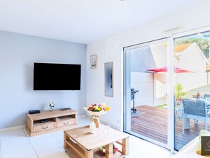 Ein modernes Wohnzimmer mit Flachbildfernseher, Holzmöbeln, einer Glastür zu einer Außenpatio und einer Blumenkomposition.