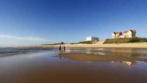 Ein Strand mit zwei Personen im Wasser, einem sandigen Ufer und zwei Gebäuden auf der rechten Seite.