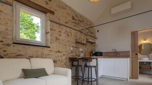Hyggelig opholdsstue med stenvæg, sofa, vindue og et køkkenområde med vask og barstole.