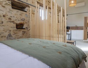 A bedroom with a stone wall, a bed with a green bedspread, a wooden partition, a kitchen area with a sink and stools, and a hanging light fixture. - Studio with AC, peaceful village (Chenillé-Champteussé)